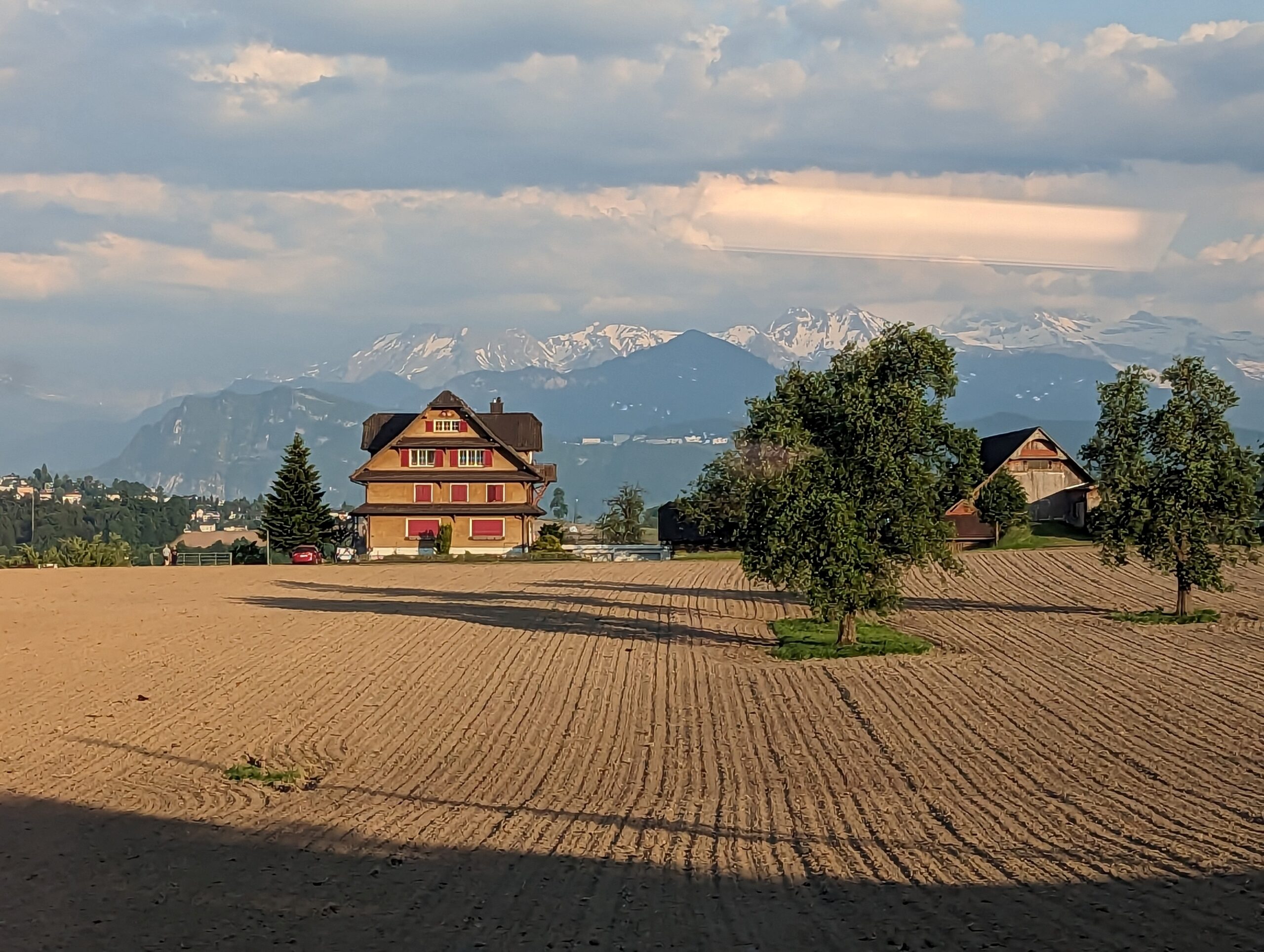 A scenic view from a train window showcasing a vast brown field with a charming chalet nestled at the foot of majestic mountains.