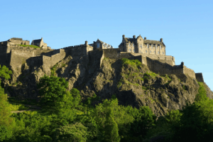 "Majestic view of Edinburgh Castle perched atop Castle Rock, with its ancient stone walls and towers set against a clear blue sky, overlooking the vibrant city of Edinburgh below."