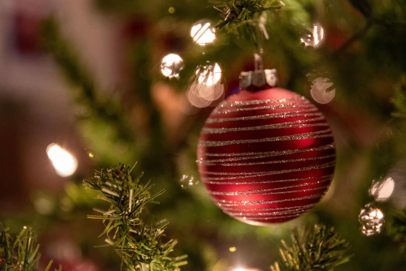 Close-up of a shiny red Christmas ornament hanging on a tree branch, with soft, blurred green needles and warm holiday lights in the background.