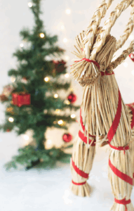 Straw Yule Goat ornament with a red ribbon standing in front of a decorated Christmas tree with warm white lights.