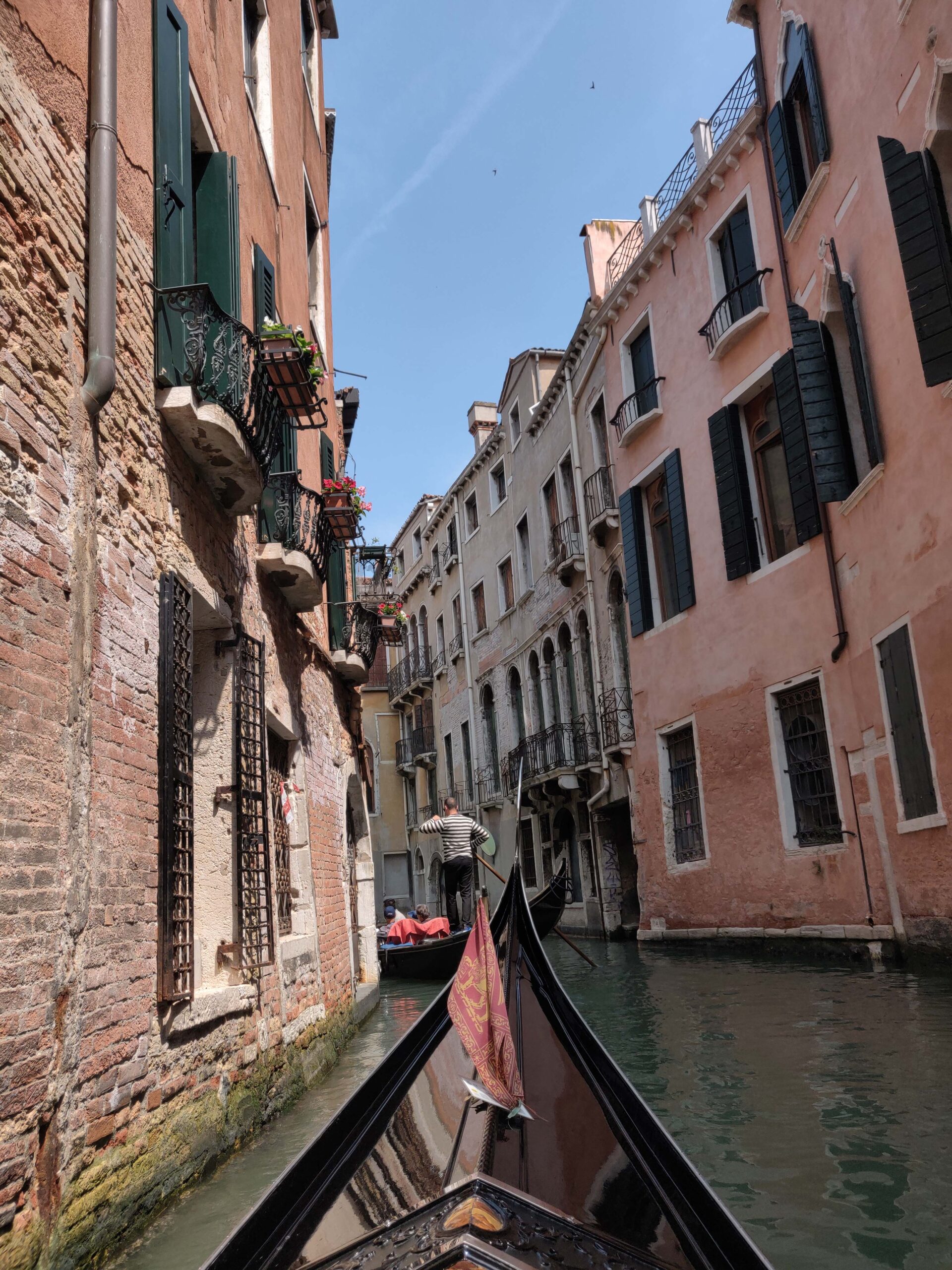 View from a gondola gliding along a narrow canal in Venice, with historic pastel buildings on both sides and small bridges arching overhead.