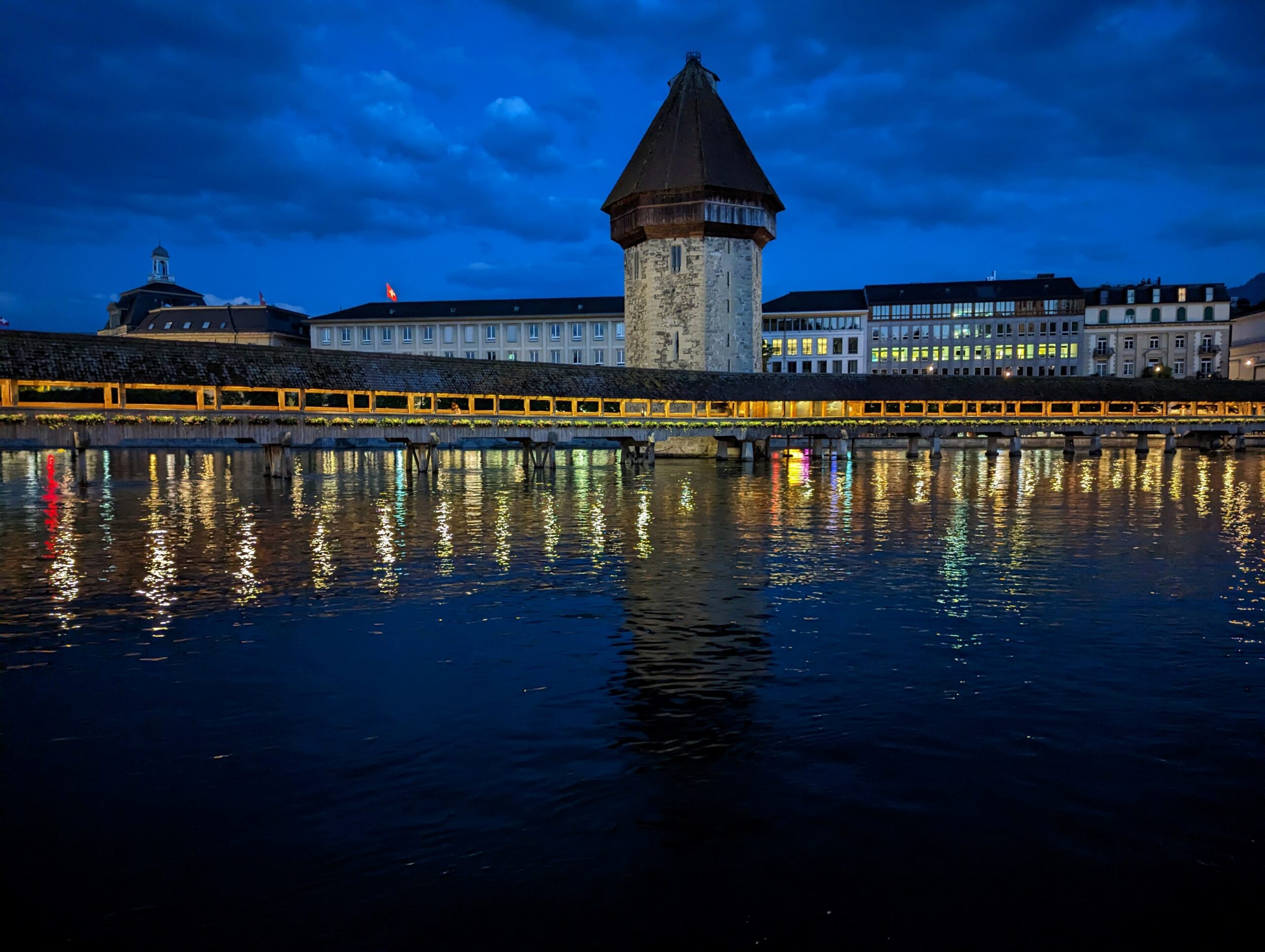 Kapellbrücke, the illuminated wooden covered bridge in Lucerne, Switzerland, reflecting in the Reuss River at night, with the Water Tower and old town buildings behind it.