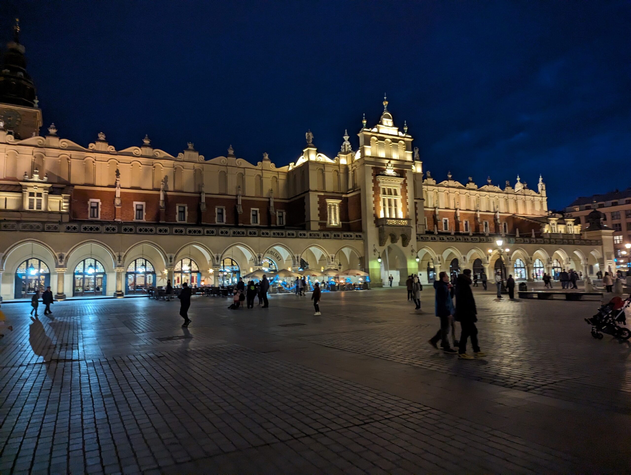 Cloth Hall in Kraków’s Main Market Square at night, warmly lit arches and historic façade glowing against a dark sky, with people walking across the cobblestone square.