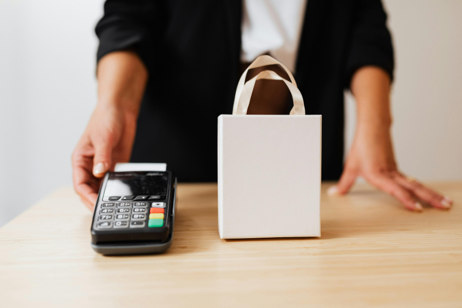 Close-up of a card payment terminal on a shop counter, with a shopping bag beside it and a store attendant standing in the background.